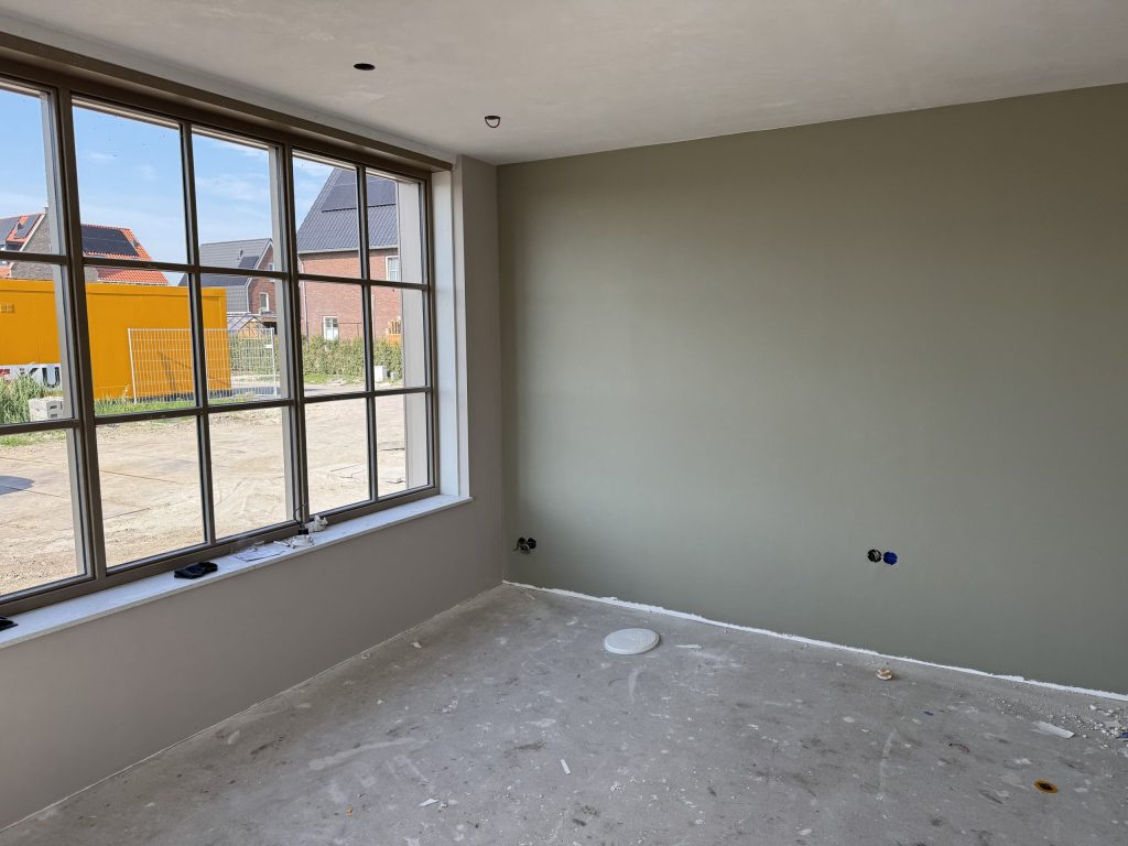 Nieuwbouwkamer met een wand in een rustige grijsgroene tint, een grote raampartij met roedes en een kale betonvloer