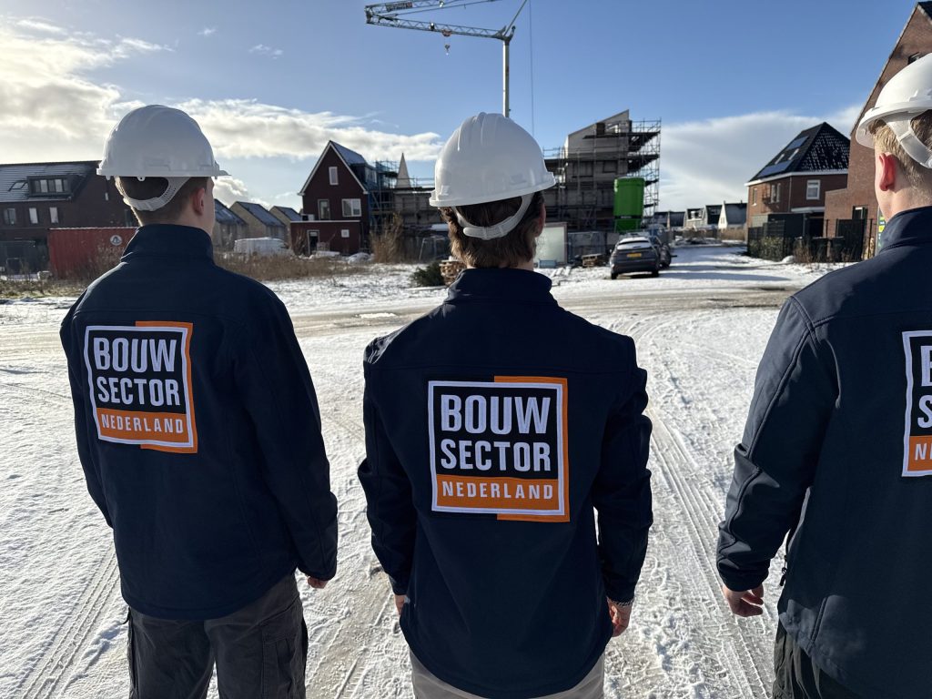 Drie vakmensen van Bouwsector Nederland met witte helm en donkerblauwe jas kijken naar nieuwbouwwoningen in aanbouw met een kraan